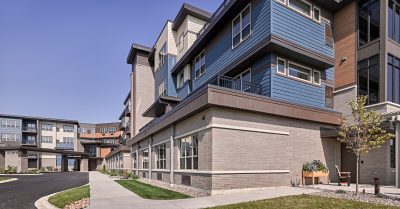 Three story senior living complex with brick and blue paneling, featuring large windows. The building has a clean, seamless design with a small, landscaped garden and a paved walkway and driveway leading into the complex.