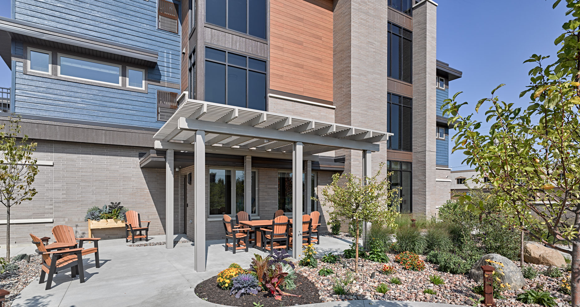 A modern multi-story building with blue and beige exterior walls features a covered outdoor seating area. The patio has wooden chairs and tables surrounded by landscaped greenery and plants, creating an inviting and peaceful atmosphere. The sky is clear and blue.