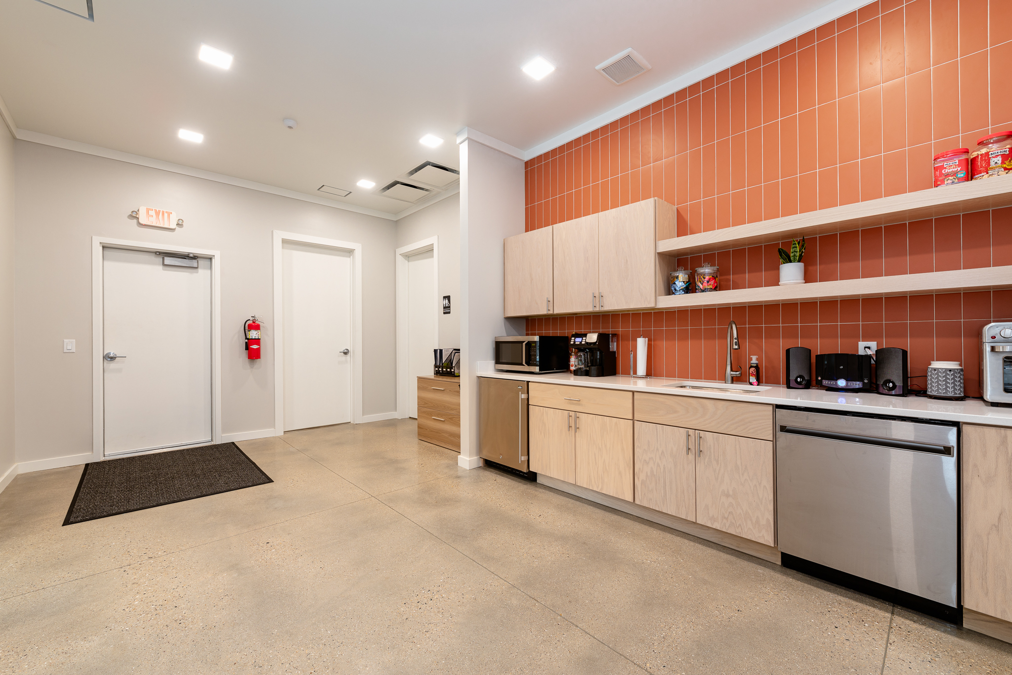 A modern office kitchenette with wooden cabinets, a stainless steel dishwasher, coffee maker, and microwave. Shelves hold plants and cups. Walls are white with an orange backsplash, and there's a fire extinguisher near two doors.