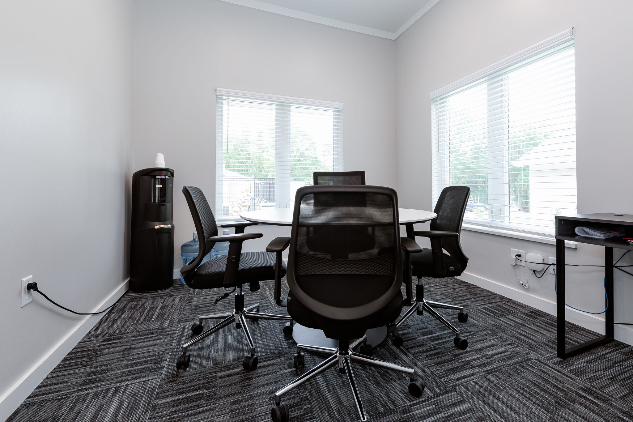 A small office meeting room features a round table with three black mesh office chairs. Large windows with blinds allow natural light to enter. A water cooler is placed in the corner, and there's a power outlet on the wall.