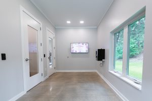 A minimalist interior hallway with polished concrete floors, two white doors, a wall-mounted screen displaying a grand opening sign, and a large window on the right overlooking greenery.