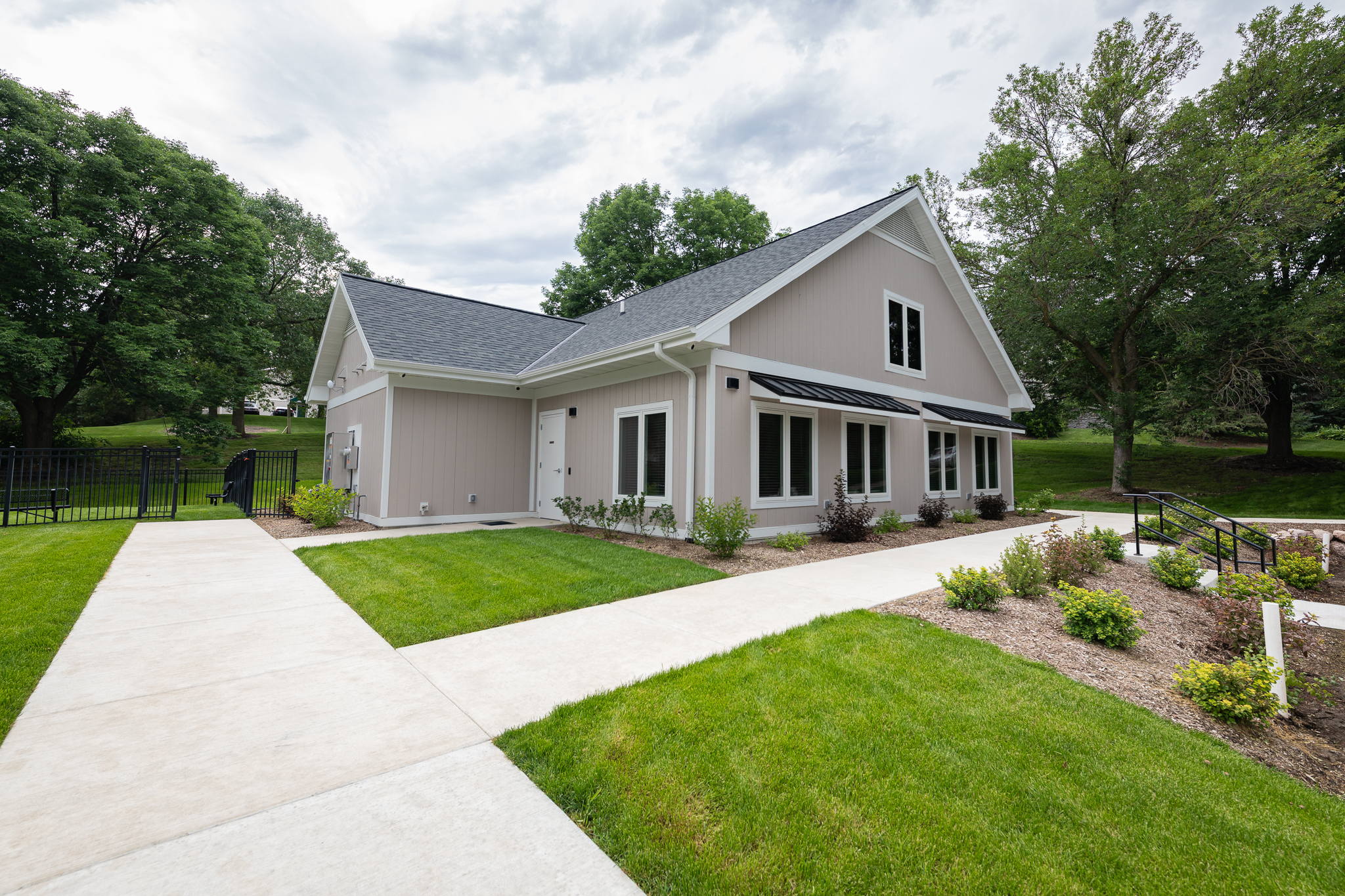 A modern, light grey house with a dark roof sits in a lush, landscaped yard. Bright green grass and neatly trimmed bushes line concrete pathways leading to the entrance. The trees in the background stand against a partly cloudy sky.