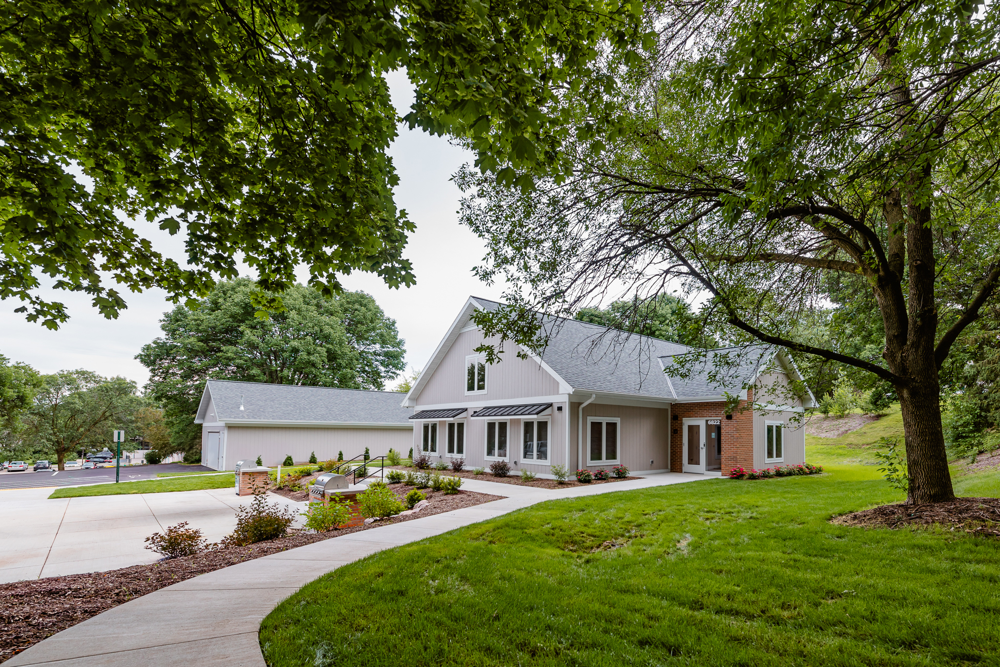 A modern white house with a gabled roof, surrounded by lush green trees and a manicured lawn. A winding pathway leads to the entrance, and there's a separate garage in the background. The scene is serene and inviting.