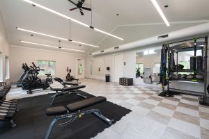 A spacious, well-lit gym with various exercise equipment, including treadmills, stationary bikes, benches, and dumbbells. A large mirror covers one wall, and a climbing rope station is set up on the right. Ceiling fans and lights are visible above.