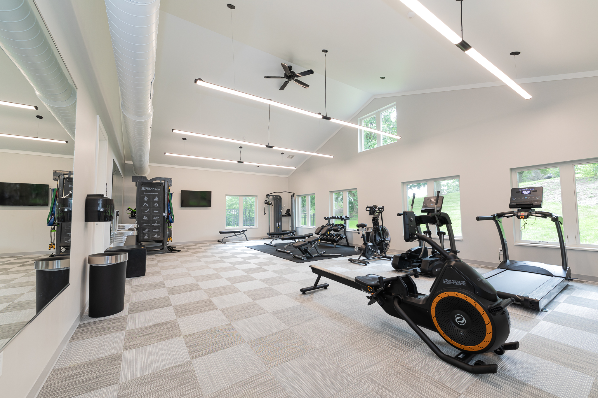 A modern gym with high ceilings, featuring treadmills, stationary bikes, and weight machines. Large windows let in natural light, and a mirror wall enhances the spacious feel. The floor is covered with a grid-patterned carpet.