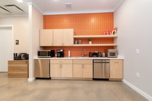 Modern office kitchenette with light wood cabinets, stainless steel appliances, and a bright orange backsplash. Includes a sink, microwave, coffee maker, toaster, and neatly organized snacks on open shelves. Neutral walls and polished concrete floor.