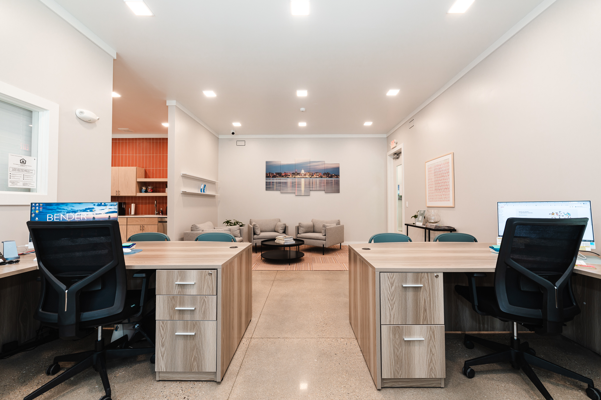 Modern office space with two wooden desks equipped with computers and black chairs. In the background, a seating area with a set of beige couches and a coffee table is visible. The walls are white with a photo and framed art hanging.