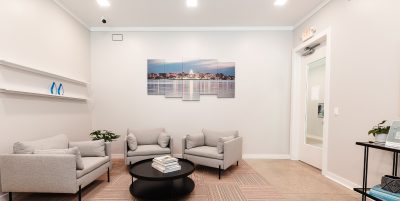 Modern office space with two wooden desks equipped with computers and black chairs. In the background, a seating area with a set of beige couches and a coffee table is visible. The walls are white with a photo and framed art hanging.