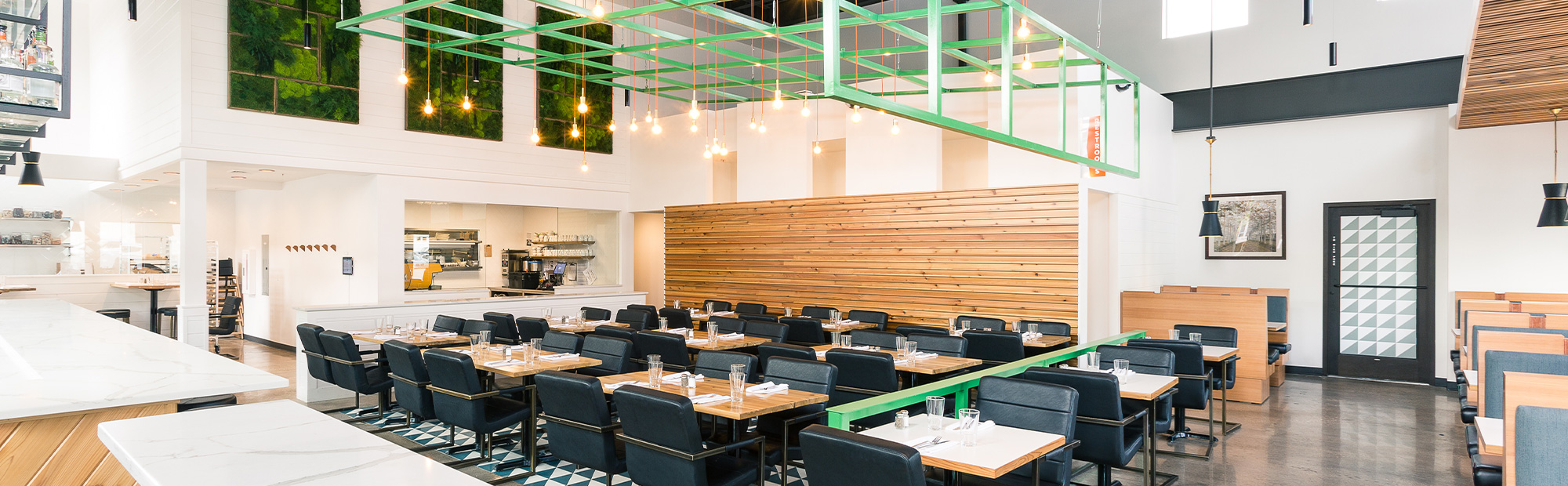 Restaurant seating area with green metal gird hanging from exposed ceiling with black chairs, concrete floors and wood table top tables