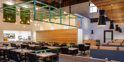 Restaurant seating area with green metal gird hanging from exposed ceiling with black chairs, concrete floors and wood table top tables