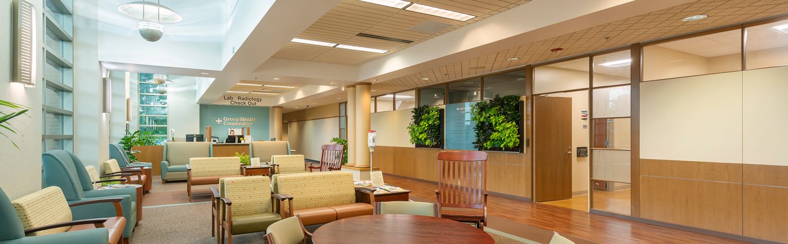 A modern waiting room with high ceilings and large windows allowing natural light. The space is furnished with green and beige armchairs, a round wooden table with chairs, and indoor plants adding a touch of nature. A reception desk is visible in the background.