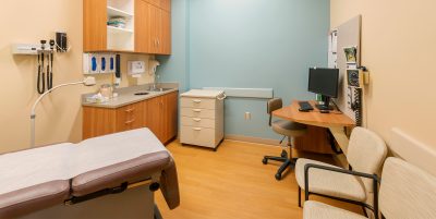 A small medical examination room with a patient bed, wooden cabinets, a beige countertop, a computer station with a chair, and medical equipment on the walls. The walls are painted light brown and teal, and the floor is wooden.