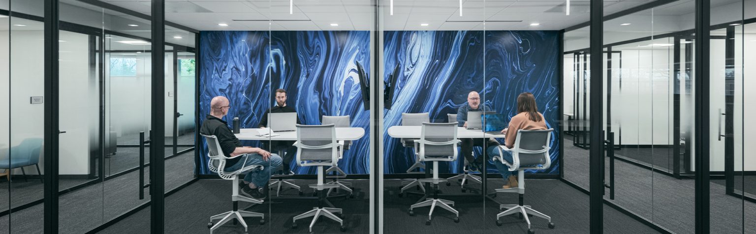 Four individuals are seated around a table in a modern conference room enclosed with glass walls. The room features a large, abstract blue and black mural on one wall. Each person is focused on their laptops or engaged in discussion.