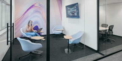 Modern office space with glass walls, showcasing a small lounge area and a separate meeting room. A person sits in the lounge on a blue chair, working on a laptop. Another person is walking down the hallway. Abstract artwork and a flat-screen TV adorn the walls.