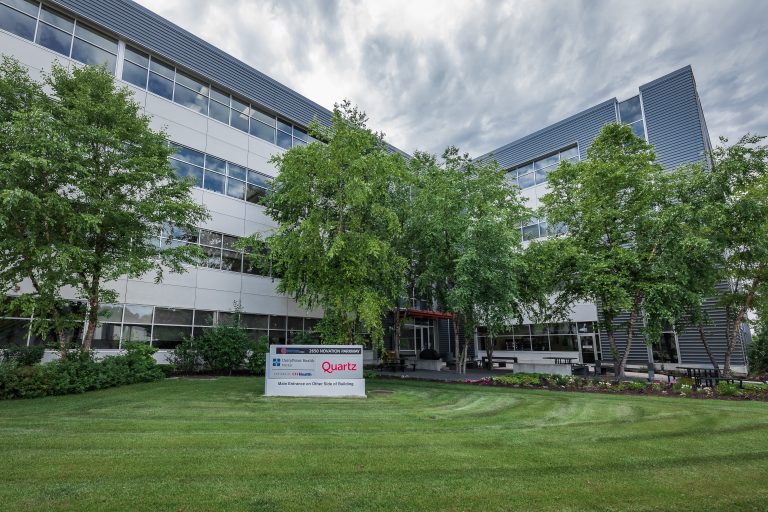 A large, modern office building with three stories, featuring glass windows and a sleek, gray exterior. Several cars are parked in the lot in front of the building, and a partly cloudy sky is overhead. Trees and greenery are around the area.