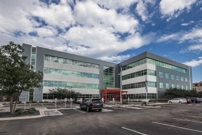 A large, modern office building with three stories, featuring glass windows and a sleek, gray exterior. Several cars are parked in the lot in front of the building, and a partly cloudy sky is overhead. The entrance has a red awning. Trees and greenery are around the area.