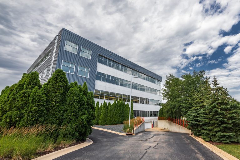 Modern four-story office building with blue and white exterior, surrounded by lush green trees and shrubs. The cloudy sky adds a dynamic backdrop to the scene.