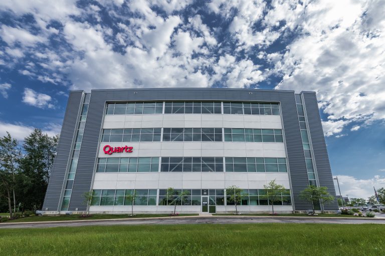 A modern glass and steel office building with the "Quartz" logo on the facade. The building is five stories high, surrounded by a grassy area and small trees. The sky is blue with scattered clouds.