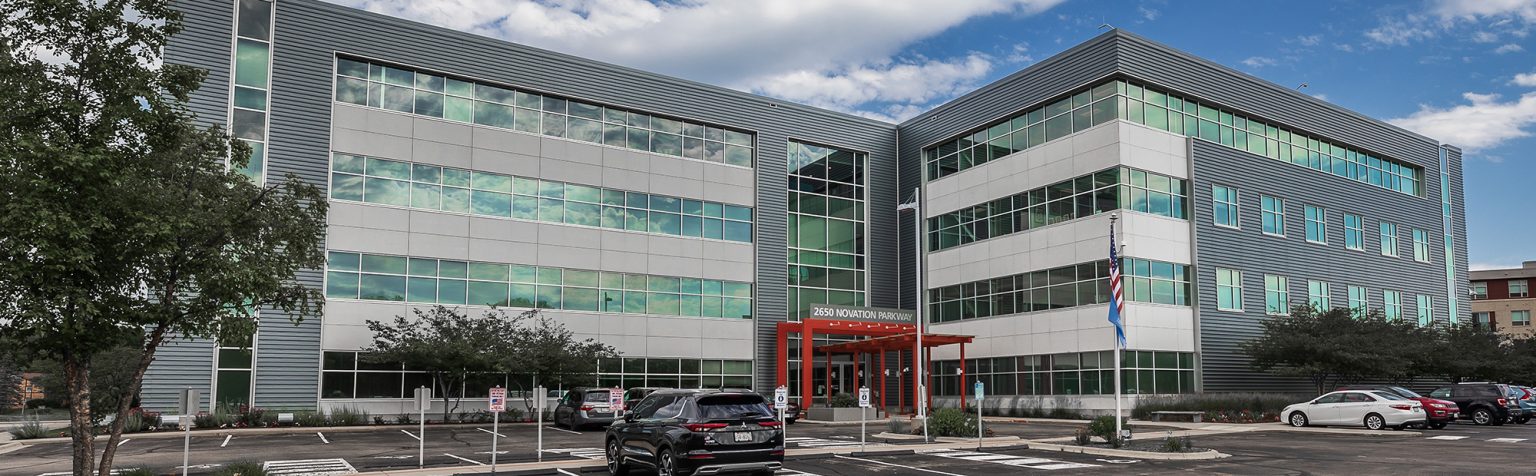 A large, modern office building with three stories, featuring glass windows and a sleek, gray exterior. Several cars are parked in the lot in front of the building, and a partly cloudy sky is overhead. The entrance has a red awning. Trees and greenery are around the area.