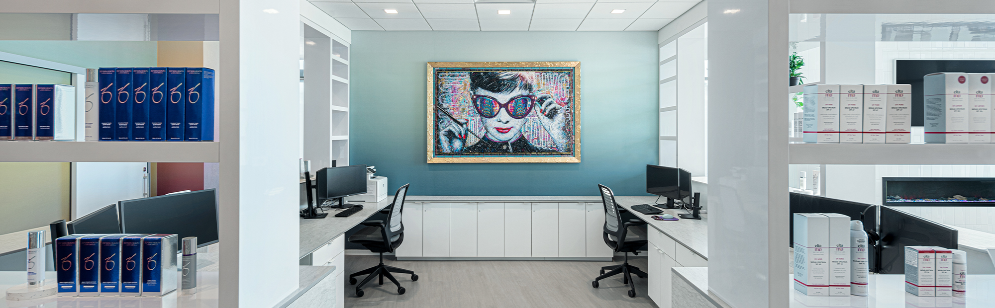 A modern office space with white shelves displaying various skincare products. Two workstations with computers face inward, and a large, colorful portrait of a woman with sunglasses is hung on the blue accent wall in the background.