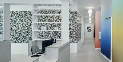 A modern, brightly lit office hallway with colorful mosaic walls and open shelving. The space includes a reception desk with a computer and chair. Hanging spherical lights line the hallway, which has doors on either side leading to different rooms.