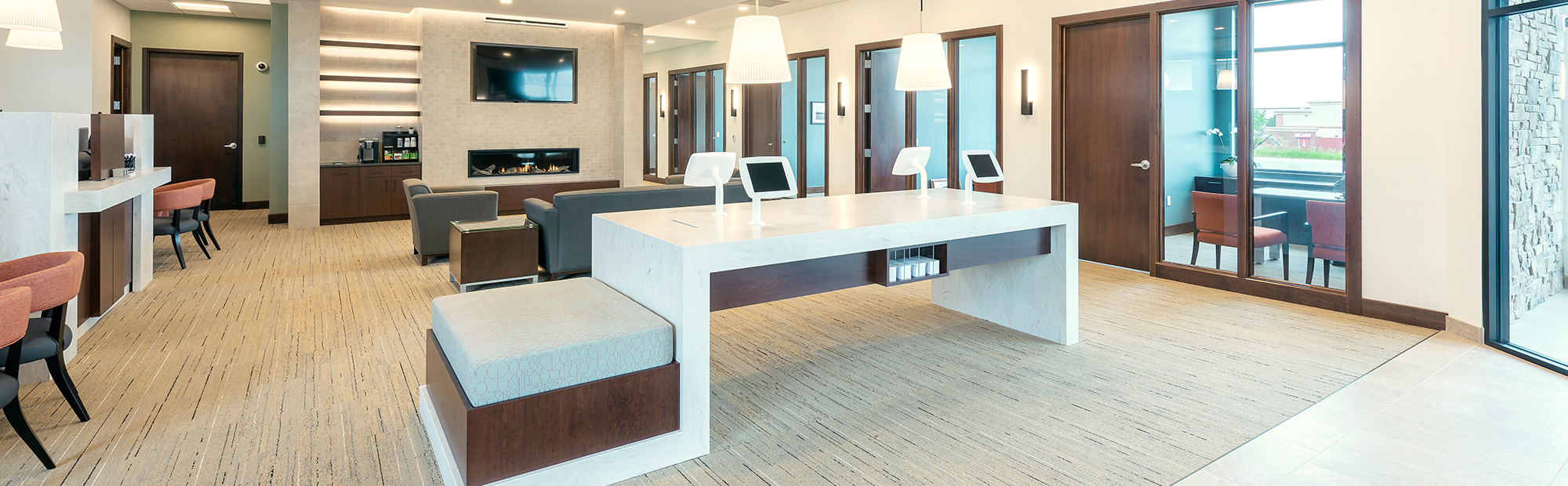 A modern design-build office space with a long white desk featuring built-in outlets and two white chairs. The room includes dark wood accents, seating areas with couches, multiple doorways lined along one wall, and a wall-mounted TV