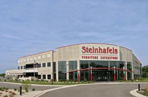 A large building with a sign reading "Steinhafels Furniture Superstore" on its facade. The structure features a modern design with expansive windows and multiple entrances. The surrounding area is landscaped with greenery and a curved driveway.