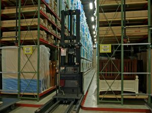 A forklift is operating in a large, well-organized warehouse aisle. Shelves on both sides are filled with boxed items and wrapped goods. The forklift is positioned centrally, ready to either pick or place items on the high shelves.