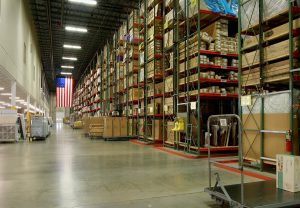 A large, organized warehouse with tall racks filled with boxes and goods stacked high. The aisle is clear with some equipment and pallets parked near the shelves. An American flag is hanging on the wall at the end of the aisle.