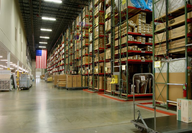 A large, organized warehouse with tall racks filled with boxes and goods stacked high. The aisle is clear with some equipment and pallets parked near the shelves. An American flag is hanging on the wall at the end of the aisle.