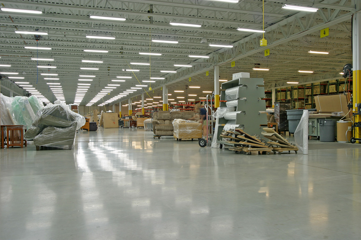 A spacious warehouse interior with high ceilings and bright fluorescent lighting. Various furniture pieces such as couches and tables are wrapped in plastic. Shelves and storage units are organized along the walls, and a cart is situated in the middle of the floor.