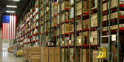 A large, organized warehouse with tall racks filled with boxes and goods stacked high. The aisle is clear with some equipment and pallets parked near the shelves. An American flag is hanging on the wall at the end of the aisle.