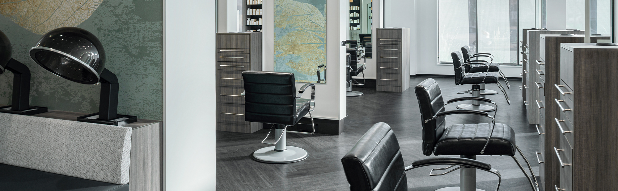 A modern and stylish hair salon interior featuring sleek black leather styling chairs, hairdryers, wood-textured counters, and large mirrors. Soft natural light filters in through large windows. Decor includes a subtle leafy mural on one wall.