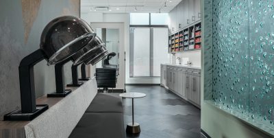 A modern hair salon with drying stations on the left, featuring dome-shaped dryers above a padded bench. To the right, there's a wall of nail polish shelves and washing stations. The room has sleek, contemporary decor with a textured glass partition.