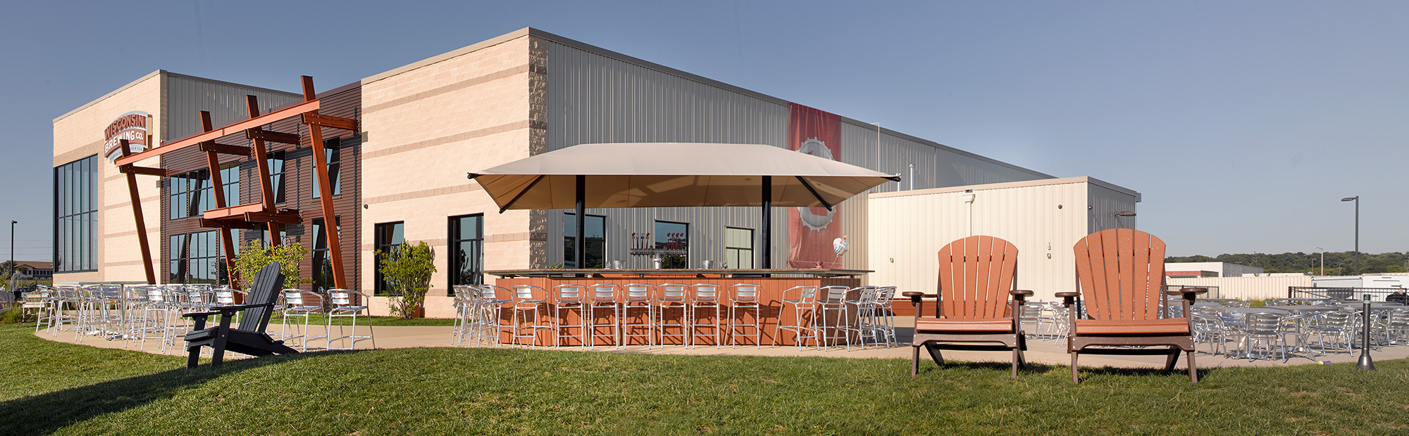 A large, modern building with a light exterior sits under a clear blue sky. An outdoor seating area with tables, chairs, and two large wooden armchairs is visible. A tall, prominent flagpole with an American flag waves in the foreground on a well-kept lawn.