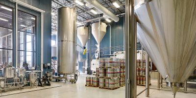 A spacious brewery with large, stainless steel fermentation tanks, kegs stacked on pallets, and various brewing equipment. The bright and clean space features high ceilings with exposed pipes and ducts, and a glass wall providing a view of adjoining rooms.