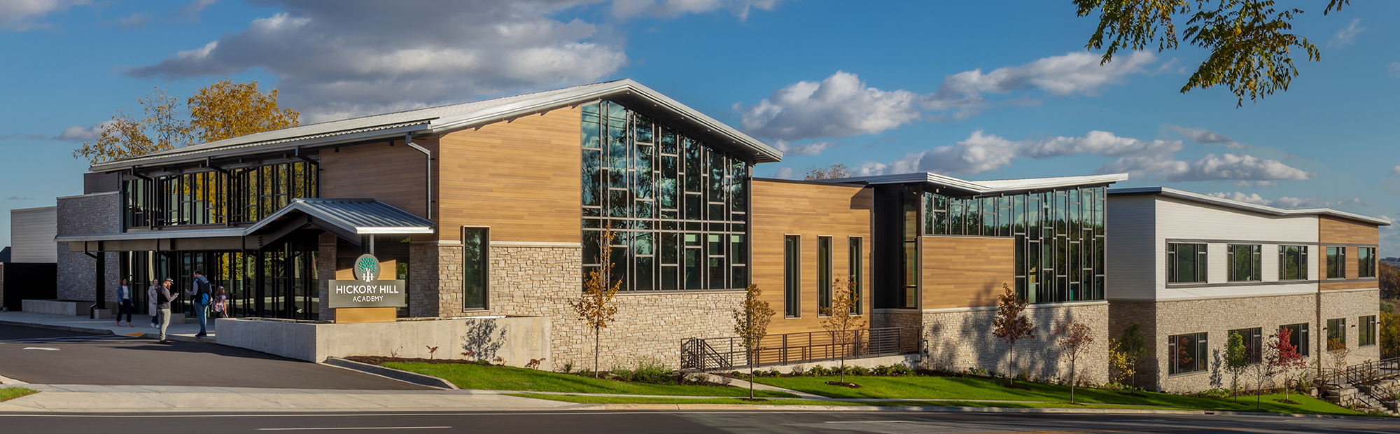 A modern building with large glass windows and a mix of wood and stone exteriors under a partly cloudy sky. The entrance has a sign for Hickory Hall Academy. People are walking near the entrance on a paved path.