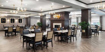 Community room with wooden floors and black-and-beige chairs around rectangular tables. There's a fireplace in the center, surrounded by dark wood panels. Elegant chandeliers hang from the ceiling, and framed art decorates the walls.