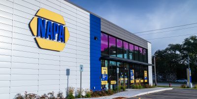 Side view of a NAPA Auto Parts store with a modern exterior featuring a large logo. The building has blue and grey panels with glass windows. A parking area with marked spaces is visible in the foreground. Trees line the background under a partly cloudy sky.