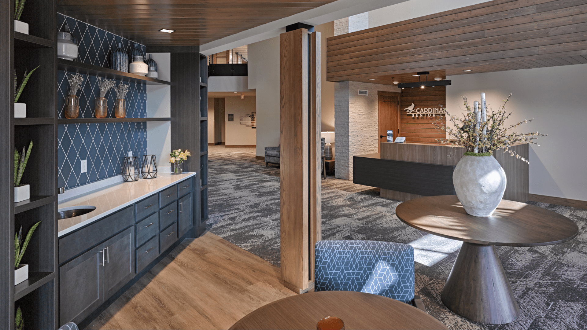 Lobby interior of a senior living community featuring a modern reception desk with a wood accent wall labeled "Cardinal Senior," a built-in beverage station with decorative shelving, and a round table with a large floral arrangement. The space is designed with warm wood tones, soft lighting, and comfortable furnishings to create a welcoming, residential feel.