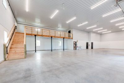 Spacious shop interior featuring a wood-railed mezzanine with stairs, steel columns, smooth concrete slab, and evenly spaced LED lights.