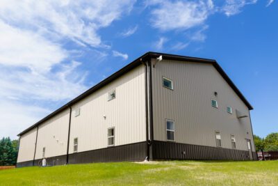Side/rear view of two-tone steel building with small windows, gutters and downspouts, set on a grassy slope under blue sky.