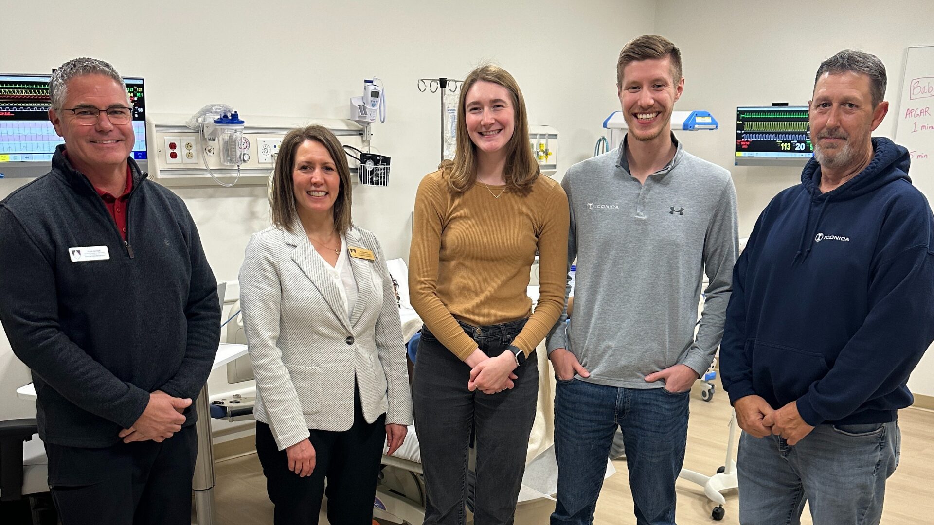 Five people stand side by side in a medical training room, smiling at the camera. Hospital monitors, wall-mounted equipment, and a bed are visible in the background, and two of the people on the right wear shirts with the Iconica logo.