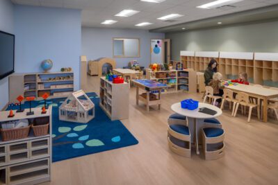 Preschool classroom with activity tables, storage, and teacher with children.