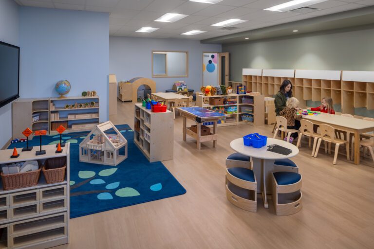 Preschool classroom with activity tables, storage, and teacher with children.