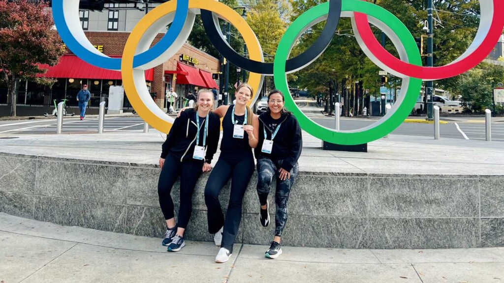 Three women wearing conference badges sit together and smile in front of the large Olympic Rings sculpture in Atlanta during an outdoor event at the AIA Women’s Leadership Summit.