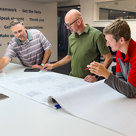 Iconica-Group-Team-Photo employees reviewing architectural blueprints on a large table in a modern office. One man in a striped polo points at the plans, a man in a green shirt writes with a pen, and a man in a red and gray jacket observes and holds a red pen. Words like 'Teamwork' and 'Get the facts' are visible on the wall behind them.