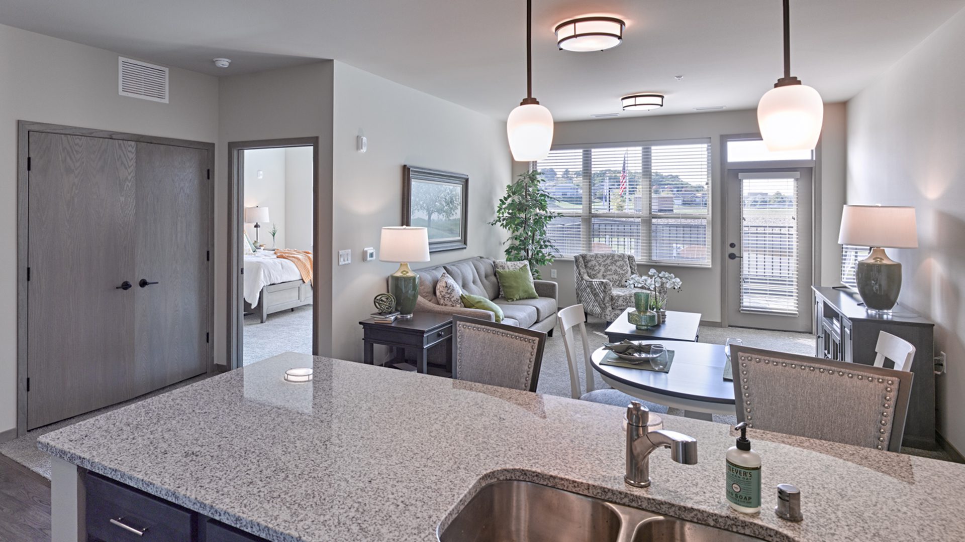 Bright, modern apartment interior featuring an open-concept kitchen with a granite countertop overlooking a dining area and living room. The living space includes a sofa, armchair, lamps, and large windows leading to a balcony. A bedroom is visible through an open doorway, and neutral tones with green accents decorate the space.