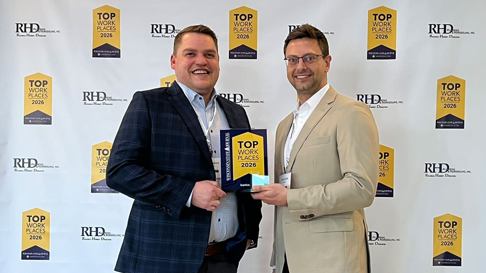 Two men in business attire stand smiling in front of a step-and-repeat backdrop with Top Workplaces 2026 and RHD logos, holding an Iconica Top Workplaces 2026 award plaque.