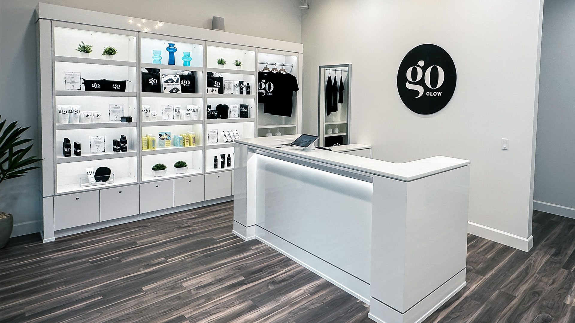 Bright, modern goGLOW reception area with a white front desk, illuminated retail shelving displaying branded products and apparel, gray walls, dark wood-look flooring, and a large round goGLOW logo mounted on the wall.